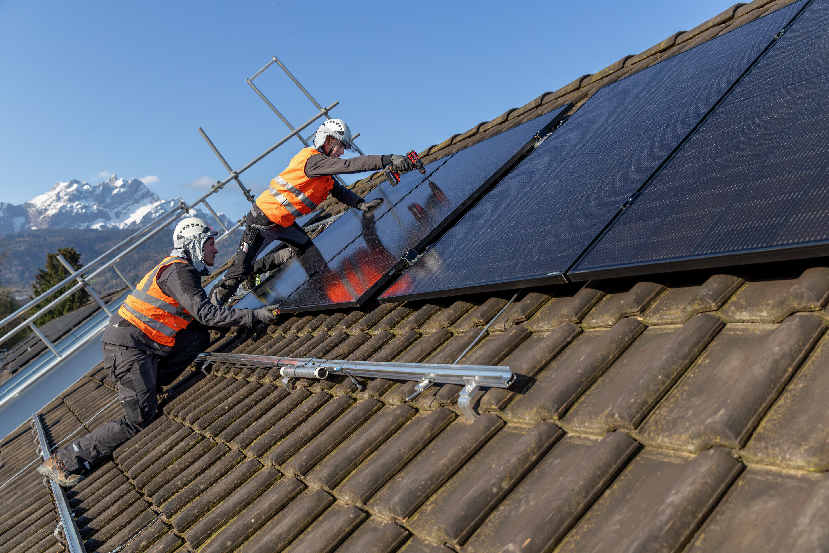 Zwei Männer mit Warnjacke und Sicherheitshelm auf einem Schrägdach mit Solaranlage.  | © Suva
