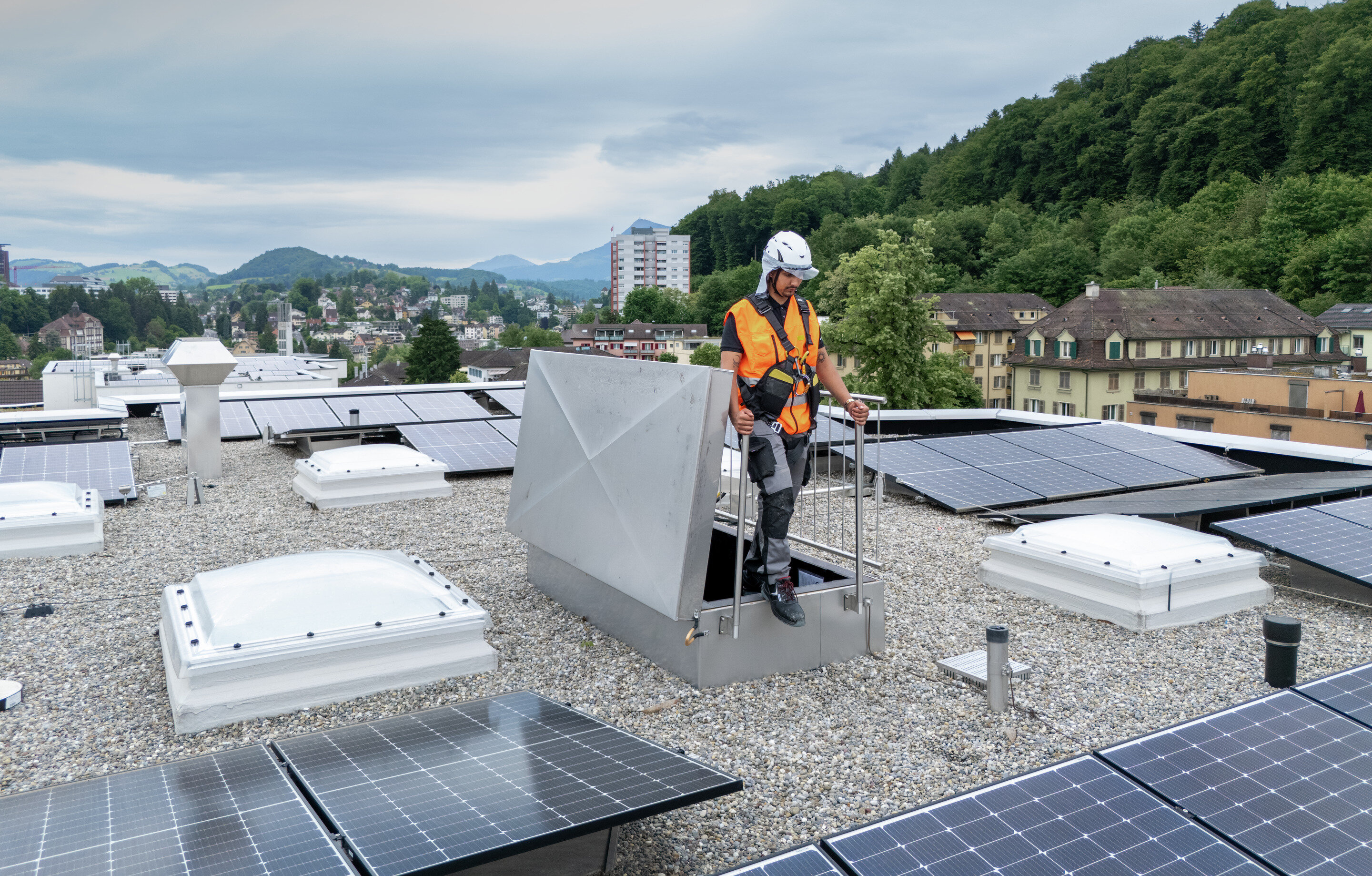 Mann mit orangener Warnjacke und Sicherheitshelm auf einem Flachdach mit Kiesboden und Solaranlage. | © Suva