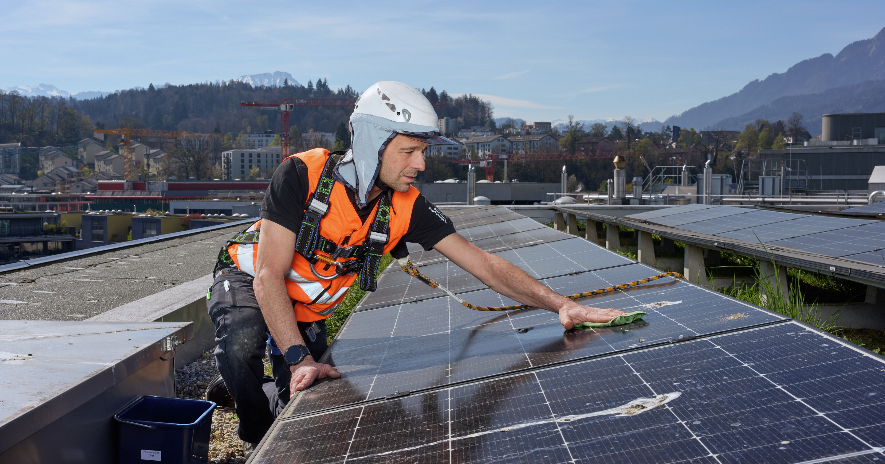Mann mit Sicherheitshelm und orangener Warnjacke befindet sich auf einem Dach mit Solaranlage. Im Hintergrund befindet sich eine Stadtkulisse und Natur.  | © Suva