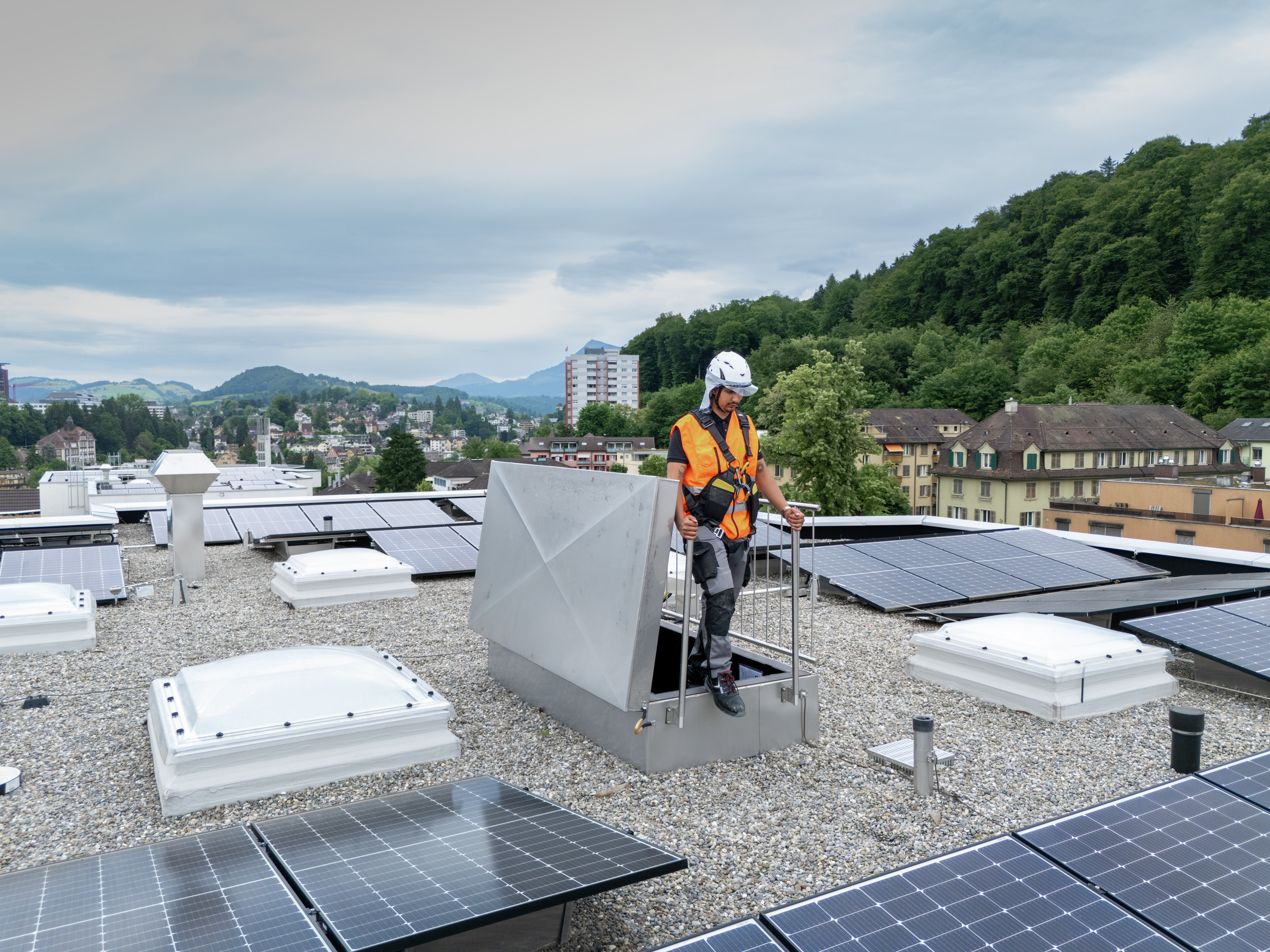 Mann mit orangener Warnjacke und Sicherheitshelm auf einem Flachdach mit Kiesboden und Solaranlage. | © Suva